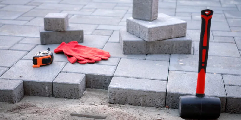 Tools and materials for brick paver installation, including a rubber mallet, measuring tape, red gloves, and gray paving stones on a sand base.
